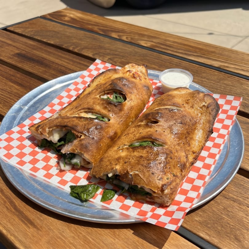 Two golden-brown calzones with visible spinach and cheese, served on a metal tray with checkered paper.