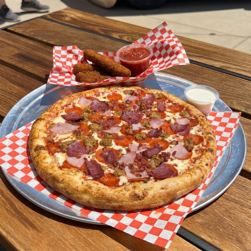 A meat lover's pizza with pepperoni, sausage, ham, and bacon, alongside mozzarella sticks and dipping sauces.